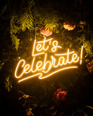 Letrero de neon amarillo con el texto "Let's Celebrate!" con un fondo decorado de vegetación verde y flores coloridas