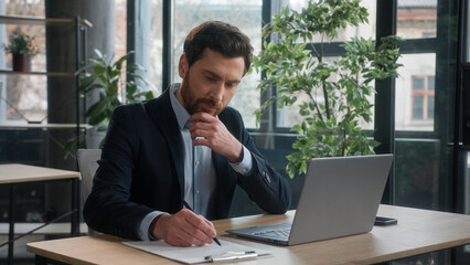 Naklejka premium Pensive puzzled Caucasian business man businessman male entrepreneur corporate employee sales manager working in office typing on laptop searching information on Internet writing notes analyzing data