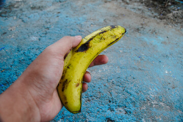 Close-up image of a slightly burnt banana held in a person's hand. The background is a textured blue surface. A slightly burnt banana held in a hand against a blue textured background