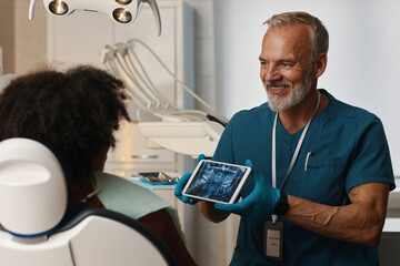 Dentist in blue scrubs showing patient digital dental X-ray on tablet. Patient seated in dental...