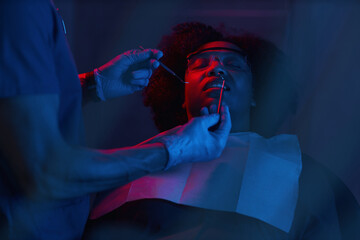 Patient lying back with eyes closed while dentist using tools to examine teeth. Scene illuminated...