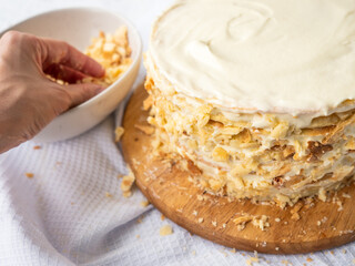 Hand sprinkling decorating crumbs over a pile of thin round layers spread with vanilla custard, bowl of dough crumbs on a white waffle towel in the blurry background. Shortcrust puff pastry Napoleon