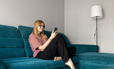 Obraz premium Beautiful blonde woman having fun online on her smartphone at home or hotel. Phone Communication and comfort concept. Smiling 30s woman daydreaming and holding mobile phone with copy space. Full body