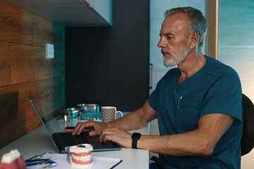 Caucasian senior dentist examining dental models while using laptop at workspace, wearing scrubs...
