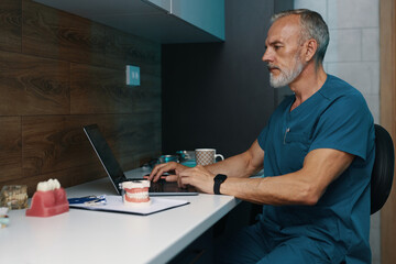 Elderly medical professional using laptop in modern office, medical tools on table. Focused on...