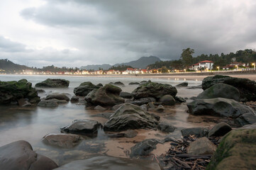 A peaceful coastal view with moss-covered rocks, calm waves, and illuminated houses along the shoreline. Cloudy sky and distant hills create a serene and natural atmosphere.