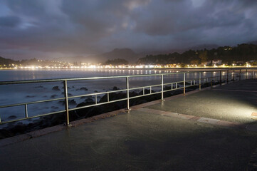 Nighttime seafront view with metal railing in the foreground, illuminated coastal town lights in the distance, and a cloudy sky. The smooth water creates a calm and atmospheric scene.