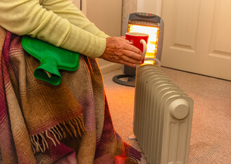 Pensioner keeping warm near oil filled and halogen heaters with hot water bottle, blanket and a warm drink