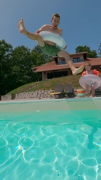 Funny excited dad and his little son jumping into a swimming pool with inflatable rings with big splashes on a sunny day, enjoying playful summer fun together. 