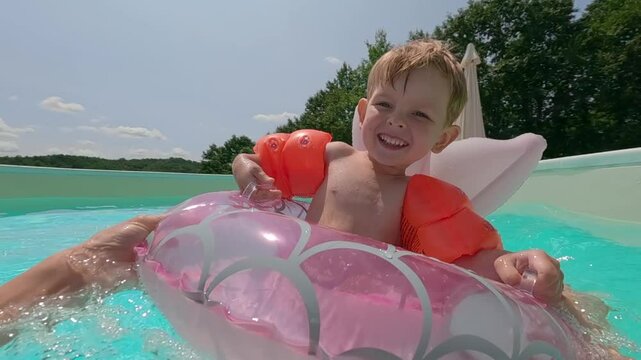 Happy child enjoying a sunny day in the pool, floating in an inflatable ring and wearing orange arm floaties, with a big smile.