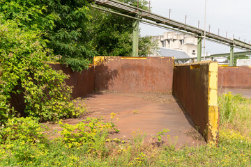 Old rusty huge open skip standing on the ground and is partially  overgrown. Concrete factory complex with pipes the background.