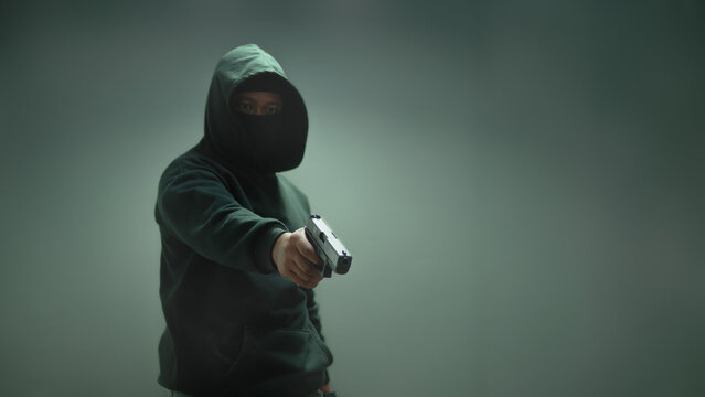 An unidentified man in a black hood and black mask stands holding a gun in various poses. Light is shining from above. Looks scary. Isolated over film studio background. Armed person.