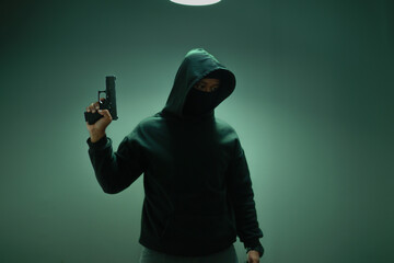 An unidentified man in a black hood and black mask stands holding a gun in various poses. Light is...