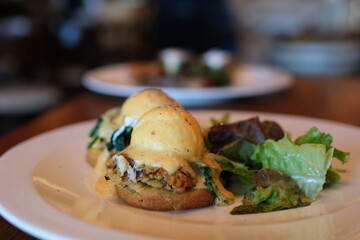 Close up of Eggs Benedict with liquid egg yolk running out. Two poached eggs on potato pancakes or toasted bread with tomatoes, fried onions and bacon with hollandaise sauce.