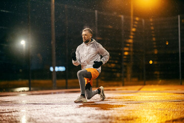 A fit sportsman is doing workouts on sports ground at night on snowy weather.