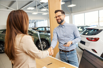 Satisfied car buyer standing at car showroom with car keys in hands and shaking hands with...