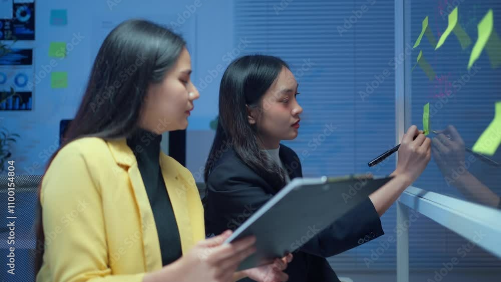 Poster two businesswomen analyzing data on sticky notes while collaborating in a modern office setting