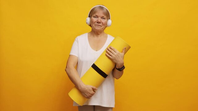 An older woman stands confidently holding a yellow yoga mat, wearing headphones and a white shirt. She radiates positivity in front of a bright yellow background, ready for her practice