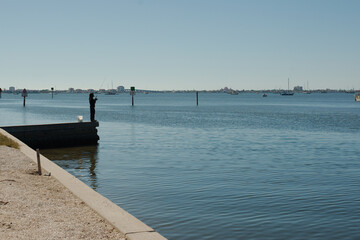 Obraz premium Leading line seawall on left man fishing off edge Gulfport, FL. View across outlet to Boca Ciega Bay from Marina. Boats in back Calm flat water. Sunny day. 