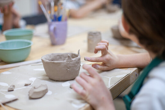 Close up of girl's hands sculpting cup with cat ears from natural clay. Children's workshop at pottery studio. Creating ceramics with own hands. Selective focus.