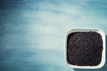 A bowl of black raw rice is placed on a blue table.The raw rice is spread out in the bowl, copy-space.