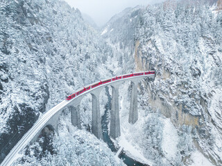 Aerial view of landwasser viaduct with a red train crossing over snowy mountains, Bergun Filisur, Switzerland.