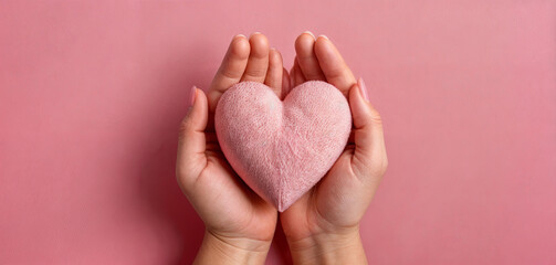 Fototapeta premium Two hands cradle a soft pink heart, set against a smooth pastel background. This gesture conveys love, warmth, and tenderness, inviting a sense of care and connection