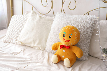Christmas gingerbread plush toy seated on bed surrounded by fluffy pillows in bright, inviting bedroom. Bed with forged iron headboard, white linen and decorative cushions, warm and cozy atmosphere