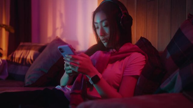 Young Asian woman in headphones is deeply engaged with her smartphone while surrounded by ambient lighting