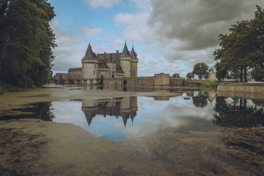 View of chateau de Sully sur Loire with reflection in the moat surrounded by trees and cloudy sky, Sully-sur-Loire, France.