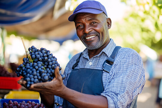 Smiling South African male farmer holds a bunch of fresh grapes at a vibrant market stall, showcasing his produce with pride and enthusiasm for the customers nearby