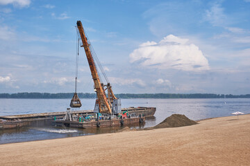 A floating crane on the calm Volga River unloads sand from a barge onto a sandy beach