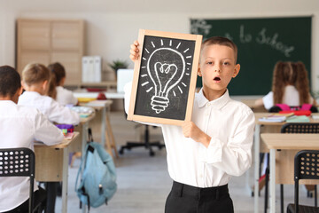 Little schoolboy holding chalkboard with drawn light bulb in classroom