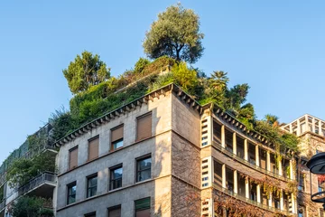  Urban building with lush rooftop garden, featuring trees and greenery cascading over the structure, blending architecture with nature under clear blue sky, Milan, Italy. Eco friendly urban environment © samael334