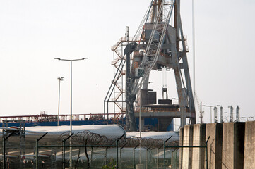 Close-up view of industrial cranes and cargo handling facilities at the port of Sines, Portugal, with a barbed wire fence in the foreground and maritime equipment in the background.