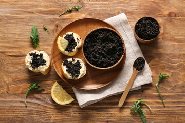 Bowls and baguette slices with delicious black caviar on wooden background