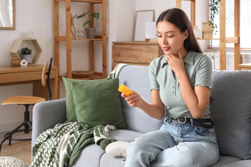 Young Asian woman with pill bottle self-examining thyroid gland on sofa at home