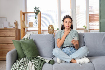 Young Asian woman in headphones with thyroid gland problem using mobile phone on sofa at home