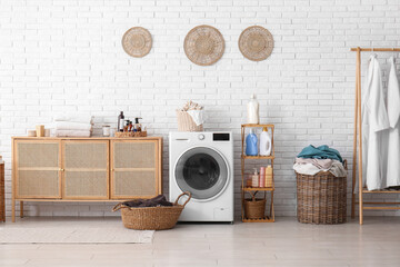 Modern washing machine, baskets, shelving unit with laundry detergent and chest of drawers in room