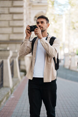A young man stands outdoors with a camera, capturing urban scenery. He is wearing casual attire and...