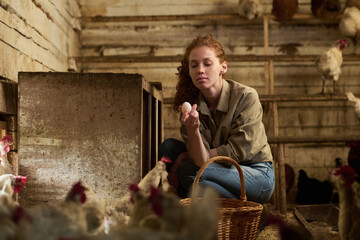 Young female farmer with fresh egg in hand sitting on squats in wooden coop for free range hens at...