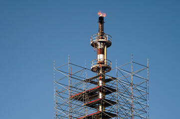 Industrial flare stack surrounded by scaffolding, with flames burning at the top against a clear blue sky, representing energy production and industrial processes.