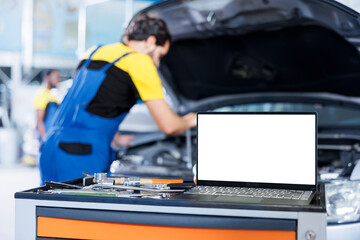 Mockup laptop on car service work station bench with engineer in blurry background replacing radiator. Isolated screen device next to certified garage employee fixing client automobile