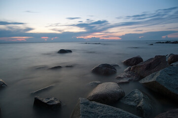 A tranquil view of the ocean at dusk, featuring smooth rocks emerging from misty waters. The soft twilight sky reflects gentle hues of blue and pink, creating a serene, atmospheric seascape.