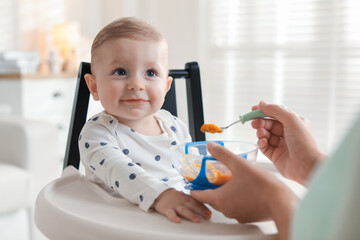 Mother feeding her cute little baby in high chair at home, closeup