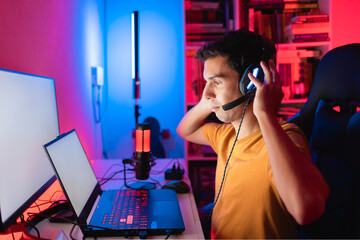 Gamer playing an online multiplayer game on a gaming laptop. He is adjusting the headset on his...