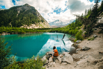 A woman is sitting on a stone near serene alpine lake with crystal-clear turquoise waters...