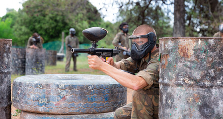 Fototapeta premium Paintball player in camouflage and mask aiming with gun in shootout outdoors