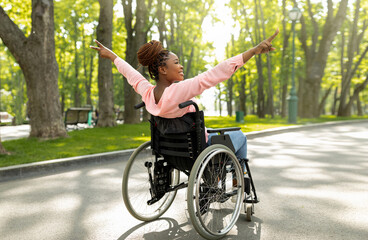 Young handicapped black woman in wheelchair on walk at green city park, showing victory gesture,...