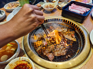 A person using chopsticks to cook meat on a sizzling barbecue grill with other dishes and condiments on the table
